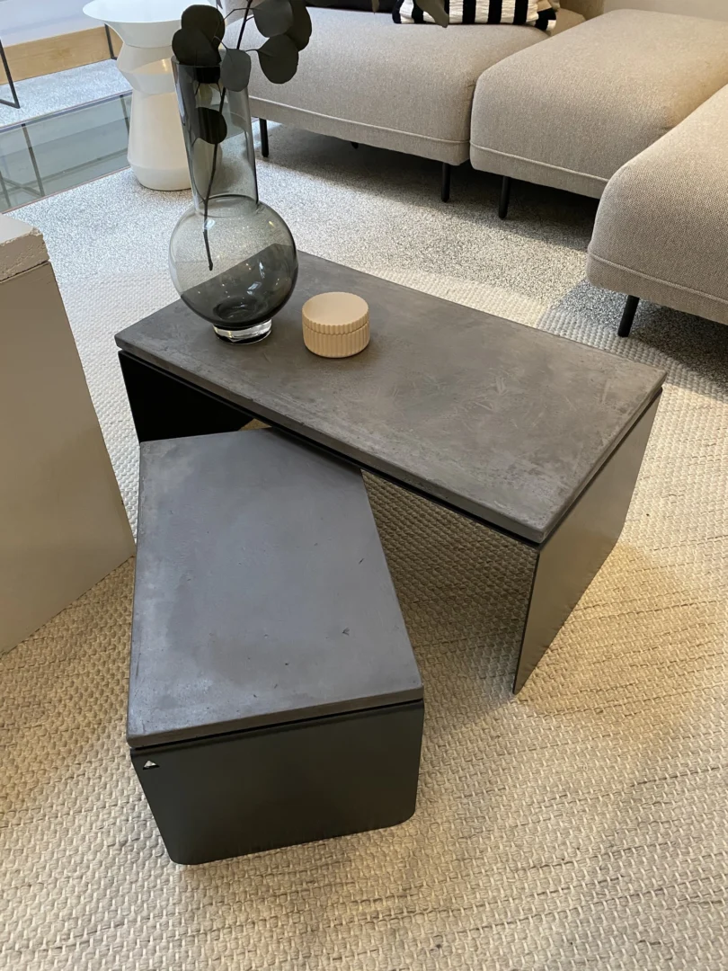 In een moderne woonkamer staat de Piramide Salontafelset met betonnen bladen - twee minimalistische, donkergrijze nesttafels op een beige vloerkleed met structuur. Op de grotere tafel staan een glazen vaas met takken en een rond, geribbeld decorstuk, naast een lichtbeige bank.