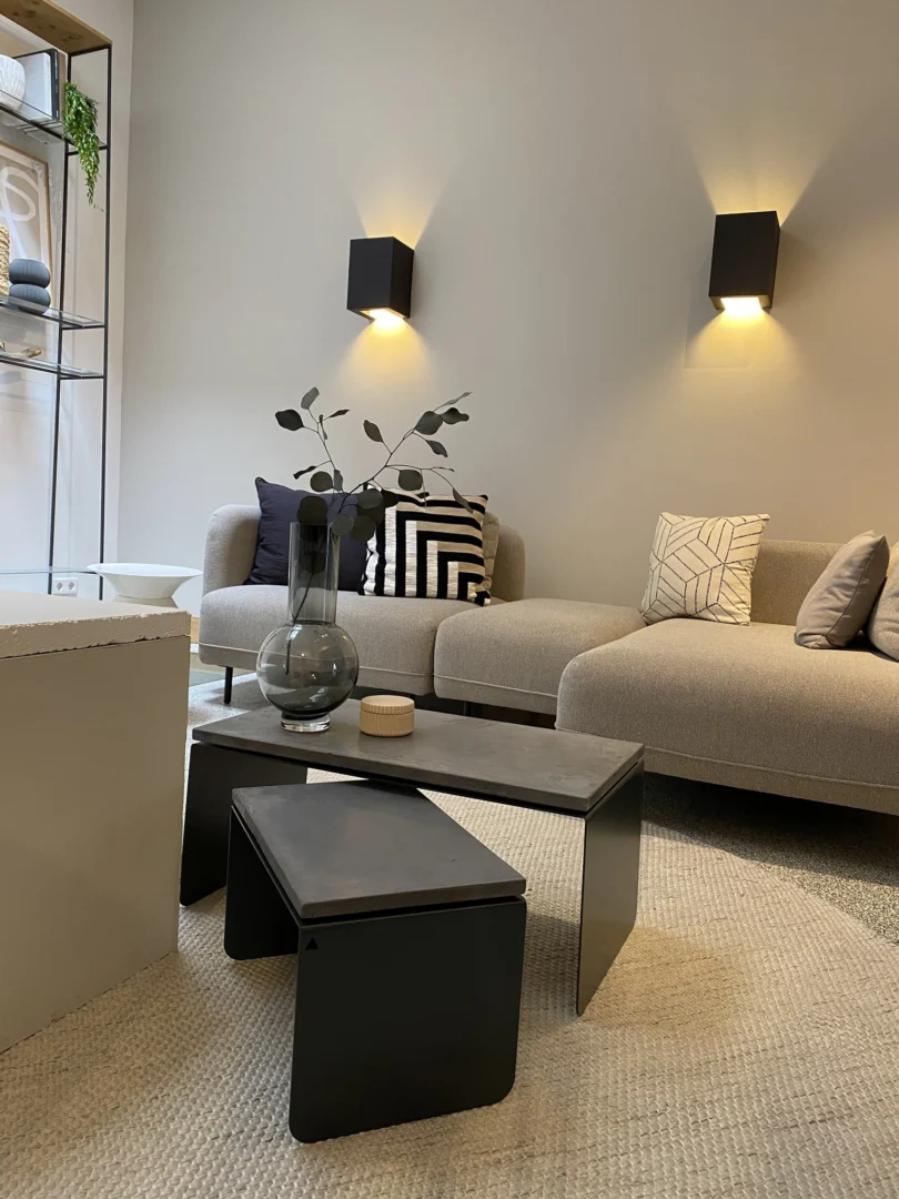 Een moderne woonkamer met een beige bank, gedessineerde kussens en een zwarte vaas met groene takken op het Piramide - Salontafelset - Betonnen blad, versterkt door zachte muurverlichting voor een gezellige sfeer.