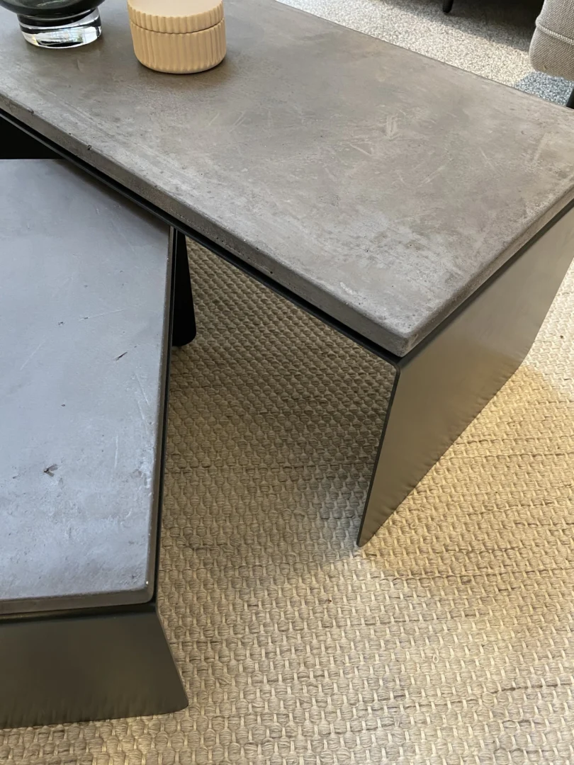De Piramide Salontafelset met betonnen bladen heeft twee moderne, overlappende rechthoekige salontafels op een beige geweven tapijt met textuur. Een ronde beige container en een glazen vaas rusten op de bovenste tafel.