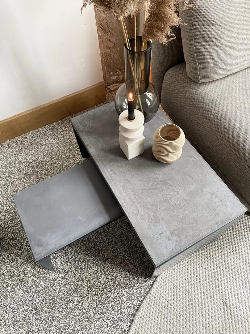 Een moderne woonkamerscène met de Piramide salontafelset met een grijs betonnen blad, met daarin een brandende kaars, een beige geribbelde vaas en een rokerige glazen vaas met gedroogd gras, naast een beige sofa op gestructureerd tapijt.