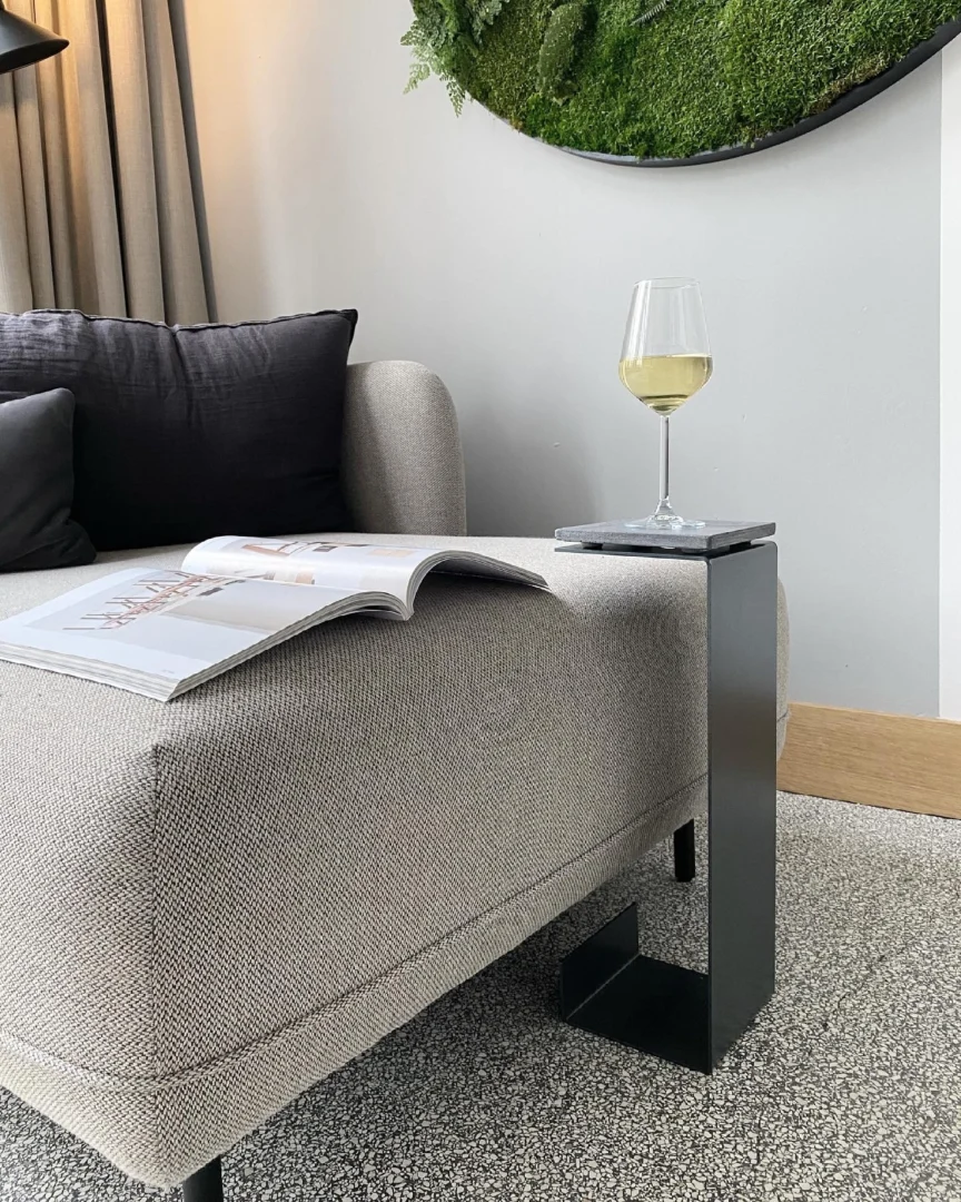 Een moderne woonkamer met een lichtgrijze bank, een donker kussen, een open tijdschrift en de Meni15 - Stalen Stand - Antraciet C-vormige zwarte bijzettafel met een glas witte wijn. Boven de bank hangt een ronde mos wanddecoratie.