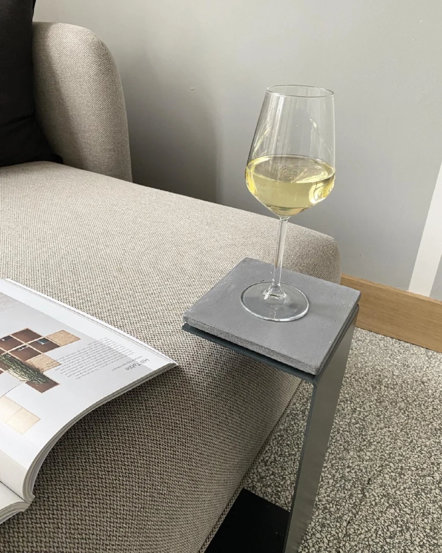 Een wijnglas staat op de Meni15 - Stalen Stand - Antraciet, gebalanceerd op de arm van een beige sofa, met een open tijdschrift in de buurt voor een moderne, gezellige setting.