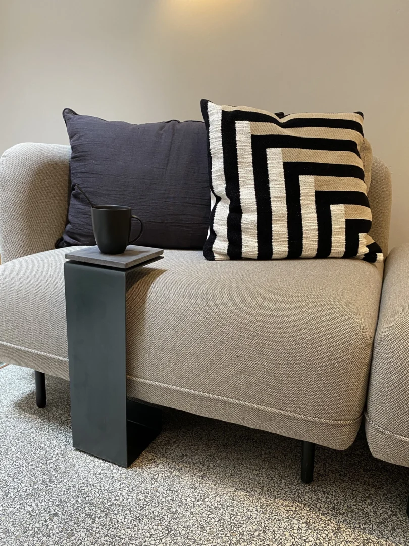 Een antraciet Meni15 Stalen Staander naast een beige sofa met twee kussens - één effen zwart, één zwart met wit geometrisch - houdt een zwarte mok vast, terwijl de sofa rust op een gespikkeld grijs tapijt.