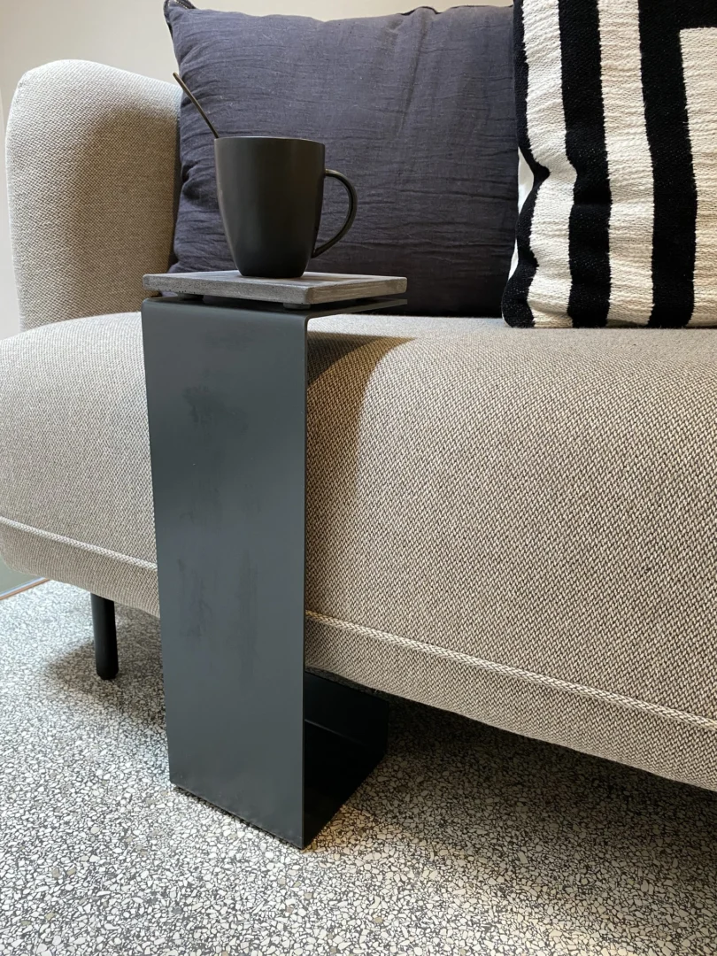 De Meni15 - Stalen Stand - Antraciet haakt over de arm van een lichtgrijze bank, met daarop een zwarte mok en lepel. Grijze en zwart-wit gestreepte kussens rusten op de bank, terwijl een gespikkelde grijze vloer de look compleet maakt.