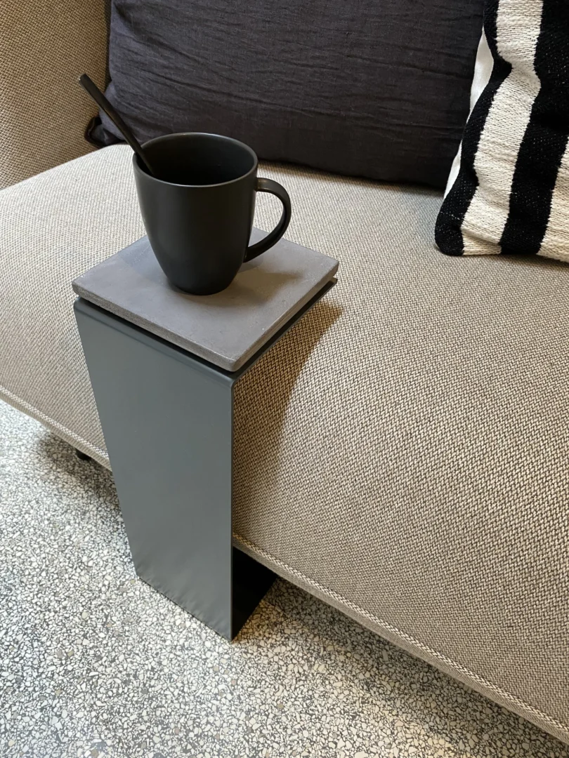 Een zwarte mok en lepel zitten op een vierkante onderzetter bovenop de Meni15 - Stalen Stand - Antraciet, een L-vormige metalen bijzettafel die over de arm van een beige bank is gehaakt, met een zwart-wit gestreept kussen op de achtergrond.