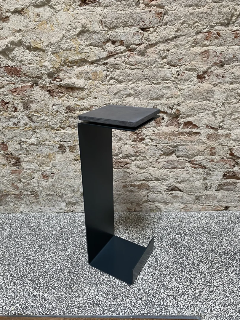 De Meni15 - Stalen Stand - Antraciet, een minimalistische zwart metalen bijzettafel met een rechthoekig ontwerp, staat op een grijs gespikkelde vloer tegen een lichte bakstenen muur met textuur.