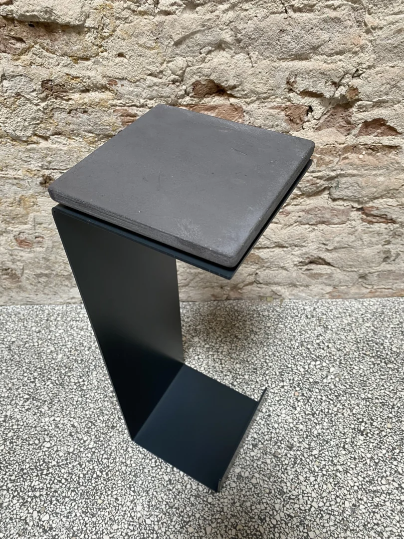 De Meni15 - Stalen Stand - Antraciet is een moderne, minimalistische zwart metalen bijzettafel met een vierkant blad en geometrisch hoekig ontwerp, afgebeeld op een gespikkelde grijze vloer tegen een ruwe beige bakstenen muur.