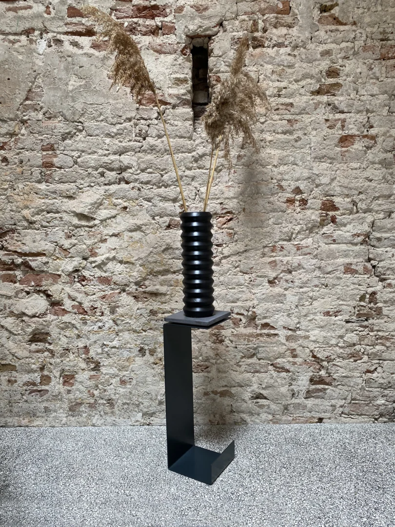 De Meni15 - Stalen Stand - Antraciet toont een geribbelde zwarte vaas met hoge gedroogde grassen tegen een gestructureerde bakstenen muur en een gespikkelde vloer.