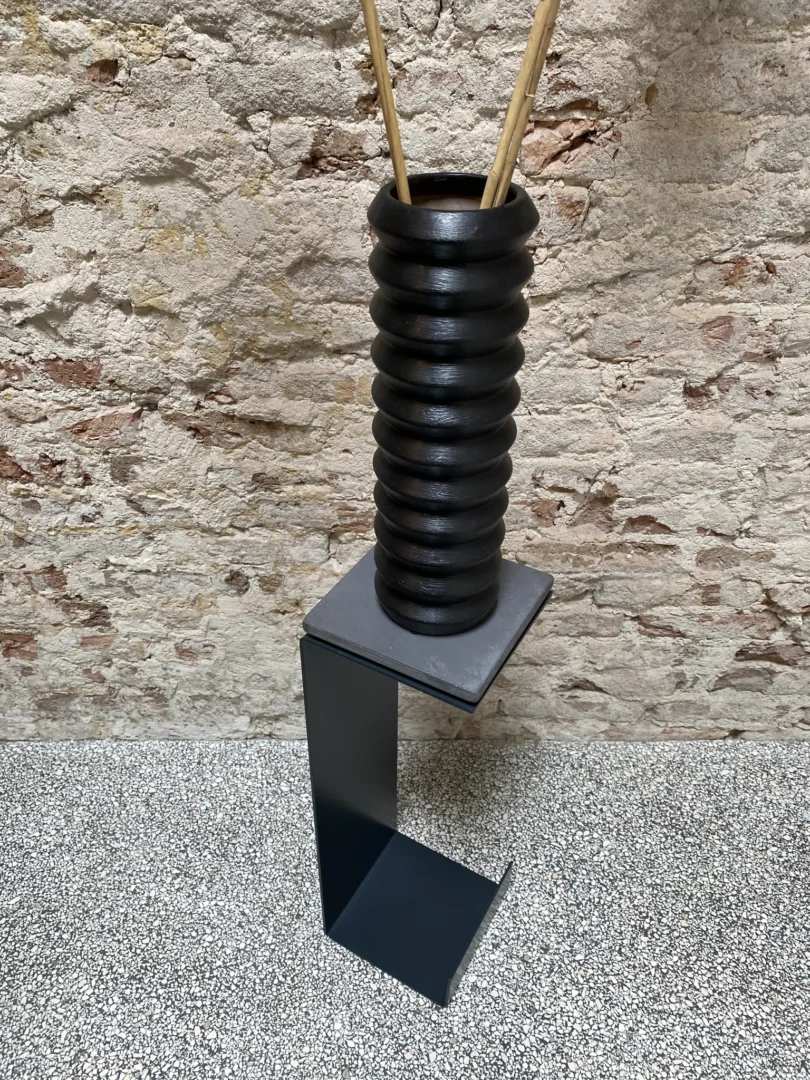 De Meni15 - Stalen Standaard - Antraciet, een kleine moderne metalen standaard met een vierkant blad en L-vormige voet, toont een zwarte geribbelde vaas met stokjes en staat tegen een muur van gestructureerd baksteen en een gespikkelde vloer.