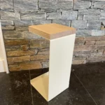 De Meni20 - Standaard heeft een modern ontwerp met een crèmekleurig metalen frame en houten blad, afgebeeld op zwarte tegels tegen een gestapelde grijze en bruine stenen muur.