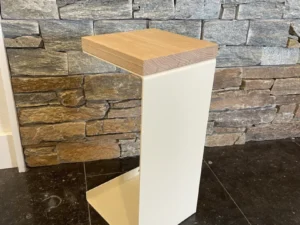 De Meni20 - Standaard heeft een modern ontwerp met een crèmekleurig metalen frame en houten blad, afgebeeld op zwarte tegels tegen een gestapelde grijze en bruine stenen muur.