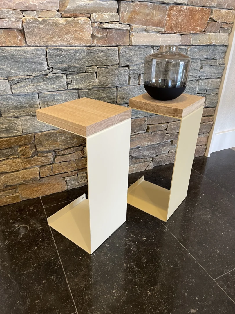 Twee Meni20 - Standaard bijzettafels met crèmekleurige metalen onderstellen en houten bovenbladen staan op een donkere vloer bij een stenen muur. Op één ervan staat een ronde glazen vaas met donkere vloeistof.
