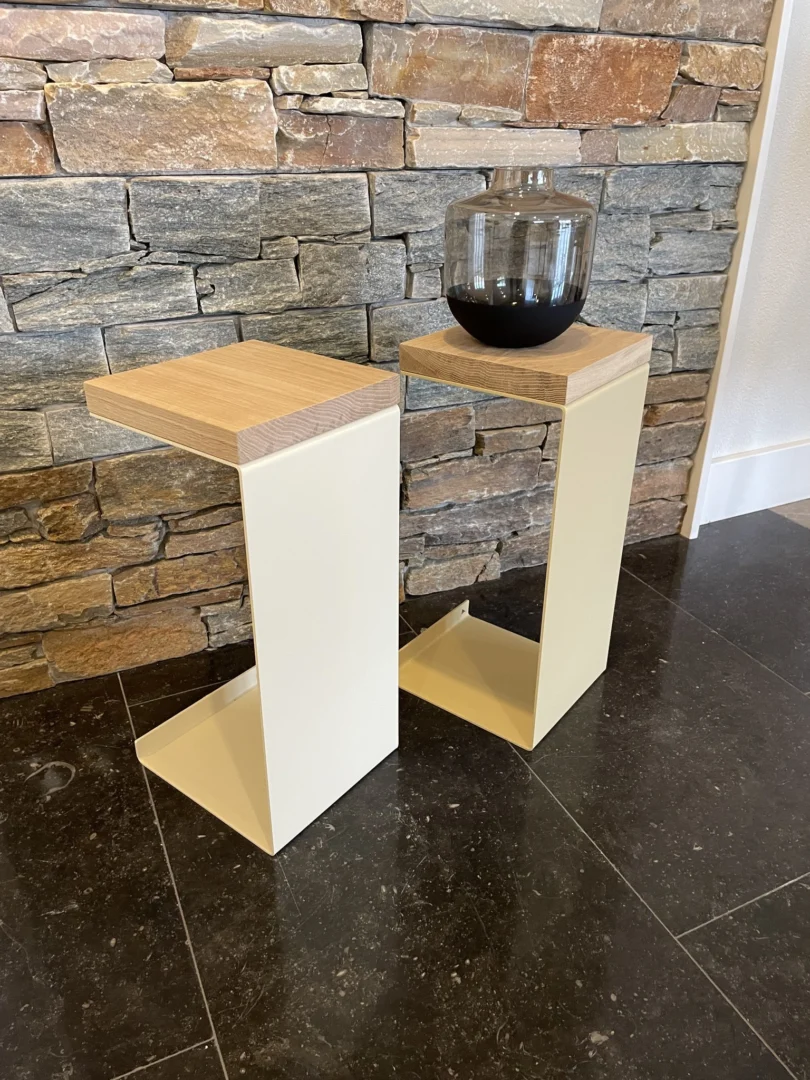 Twee Meni20 - Standaard - Crème bijzettafeltjes met houten bladen staan op een zwarte tegelvloer voor een stenen muur; op één ervan staat een transparante glazen vaas met donker water.