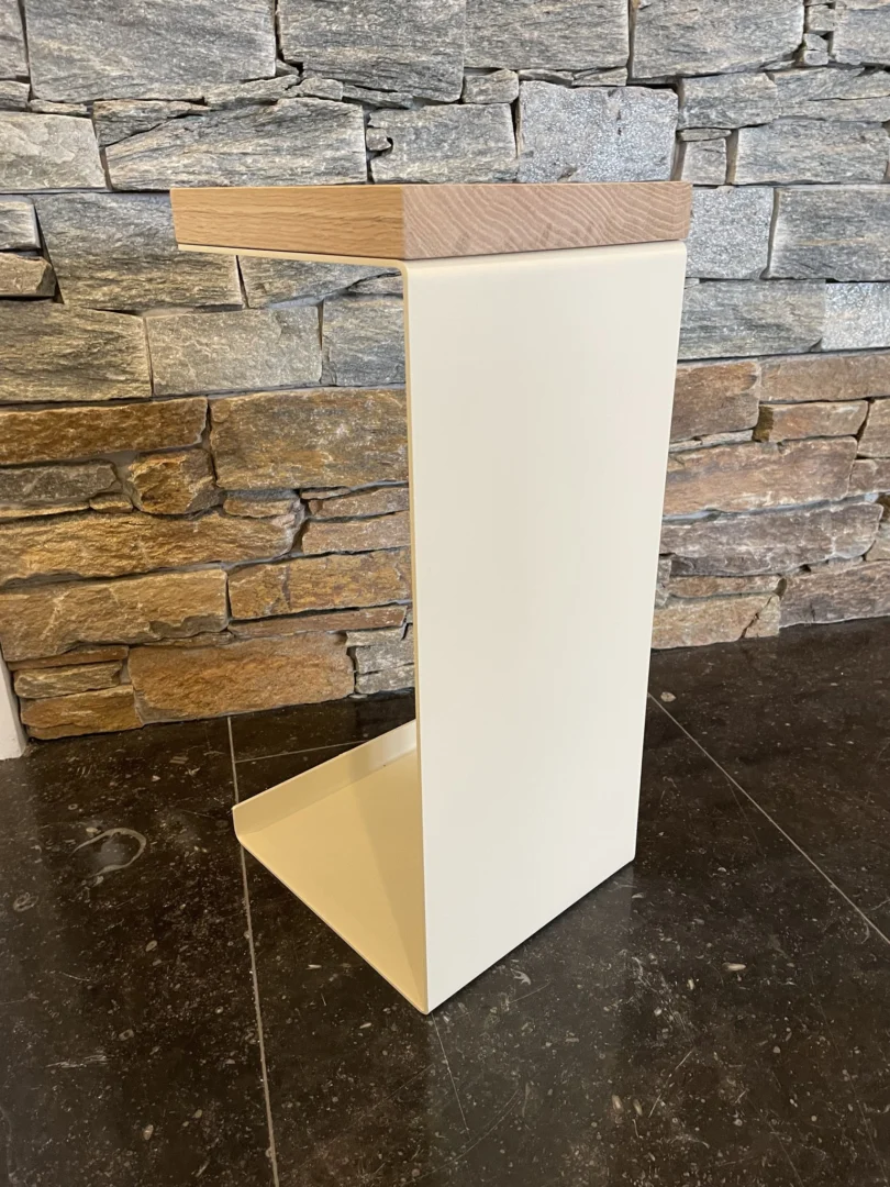 De Meni20 - Standaard heeft een minimalistische C-vorm met een crèmekleurige metalen voet en een licht houten blad, getoond op een donkere stenen vloer tegen een gestapelde stenen muur.