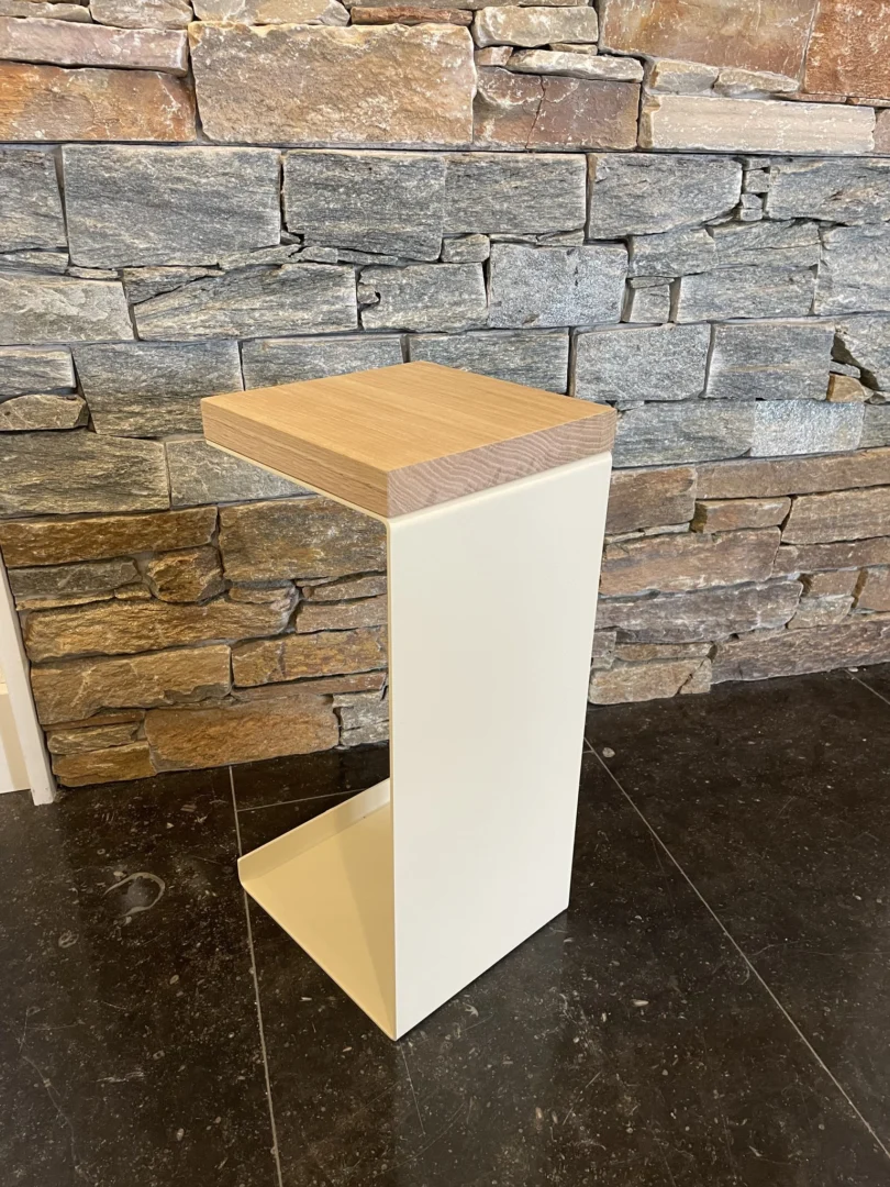 De Meni20 - Standaard heeft een minimalistisch ontwerp met een crèmekleurig, C-vormig onderstel en een houten blad, getoond op een vloer van donker steen voor een muur van gestructureerd steen.