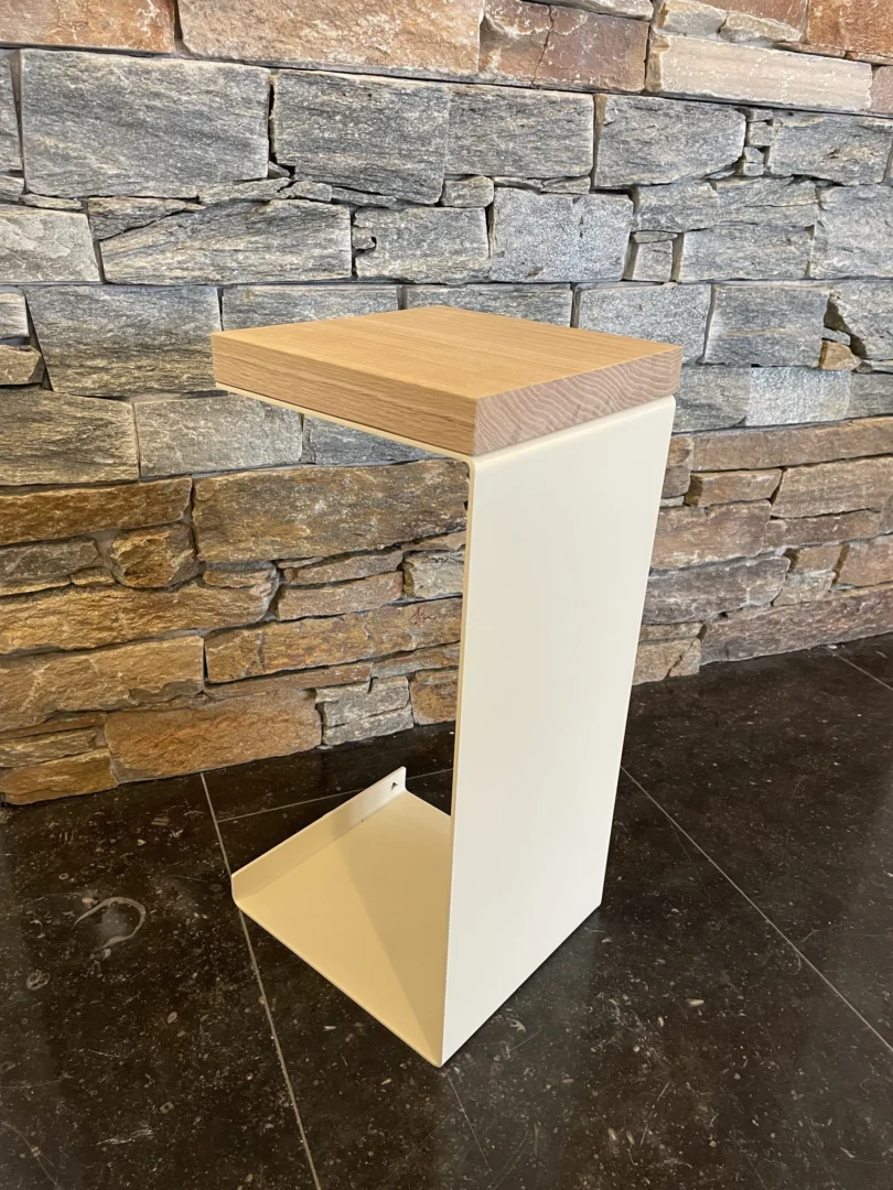 De Meni20 - Standaard - Crème bijzettafel, met een licht houten blad en crèmekleurig metalen onderstel in een zigzagontwerp, staat op een donkere tegelvloer voor een stenen muur met structuur.