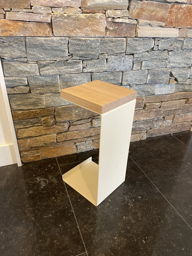 De Meni20 - Standaard heeft een minimalistisch Z-vormig ontwerp met een crèmekleurig metalen onderstel en een houten blad, dat opvalt op een donkere stenen vloer tegen een rustieke stenen muur.