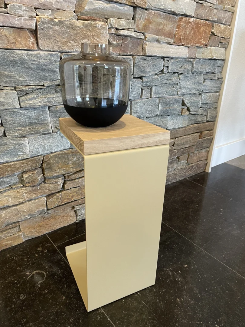 De Meni20 - Standaard - Zand bijzettafel met een licht houten blad en beige metalen onderstel toont een glazen vaas met donkere vloeistof, voor een stenen muur op een donkere tegelvloer.