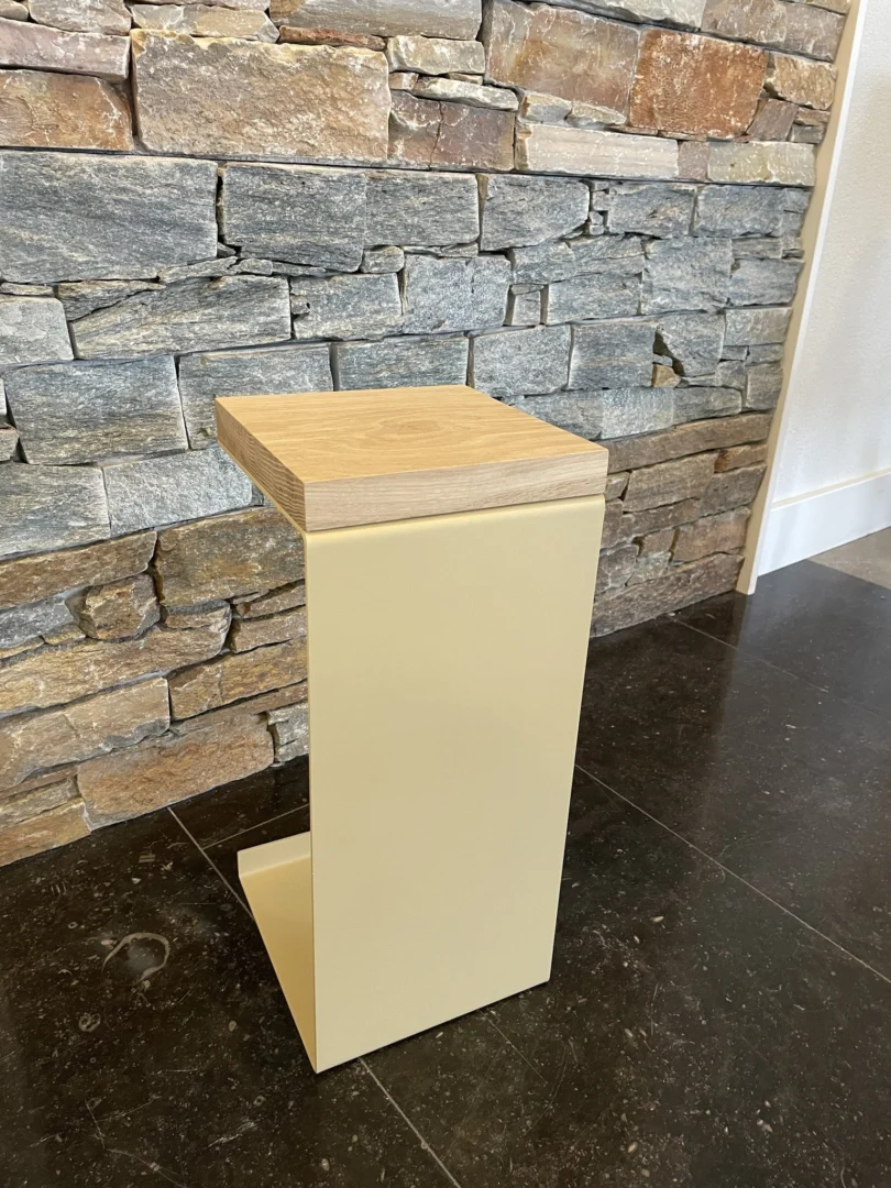 De Meni20 - Standaard - Zand kruk, met een minimalistisch beige metalen frame en lichte houten zitting, staat op een donkere tegelvloer voor een stenen muur.