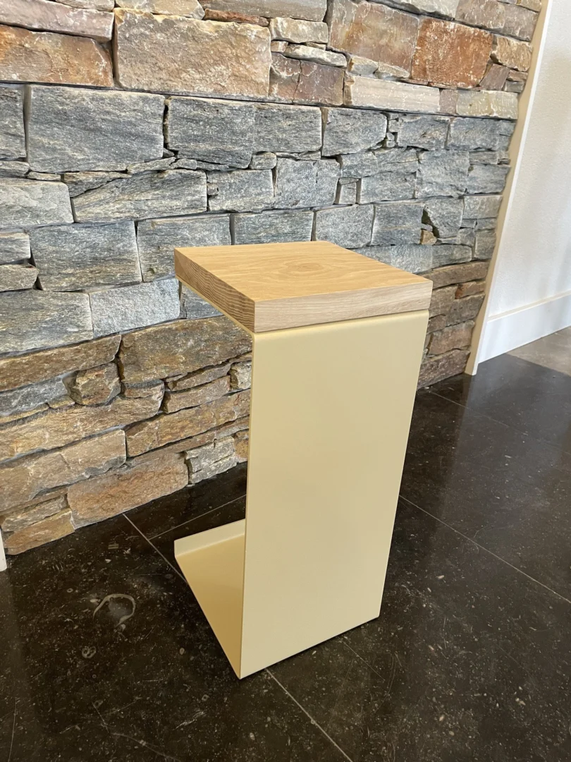 De Meni20 - Standaard bijzettafel heeft een minimalistisch ontwerp met een zandkleurig metalen onderstel in een C-vorm en een houten blad, getoond op een donkere tegelvloer voor een stenen muur.