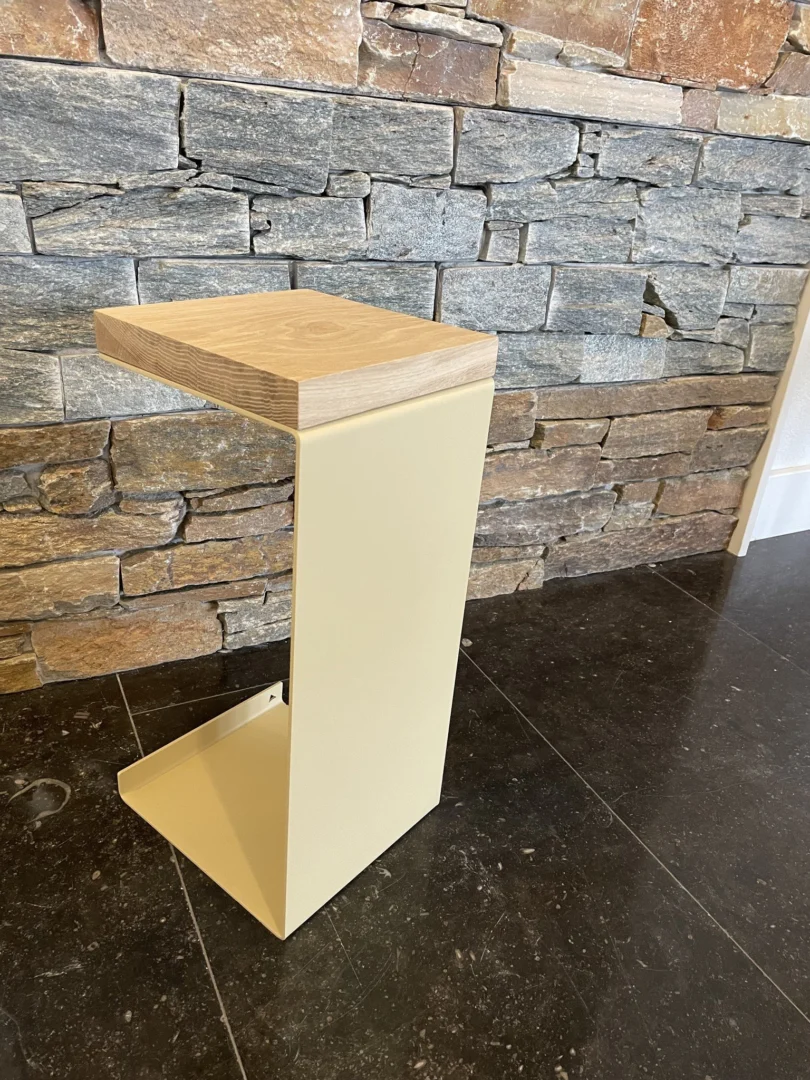 Het Meni20 - Standaard - Zand - Houten blad is een moderne, minimalistische bijzettafel met een licht houten blad en een beige metalen onderstel in de vorm van een zijwaartse Z, getoond op donkere tegels voor een stenen muur.