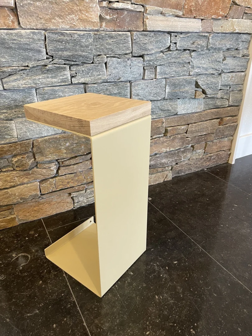 De Meni20 - Standaard - Zand heeft een houten blad en een lichtbeige hoekig metalen onderstel, dat op een donkere stenen vloer staat voor een muur van structuursteen.