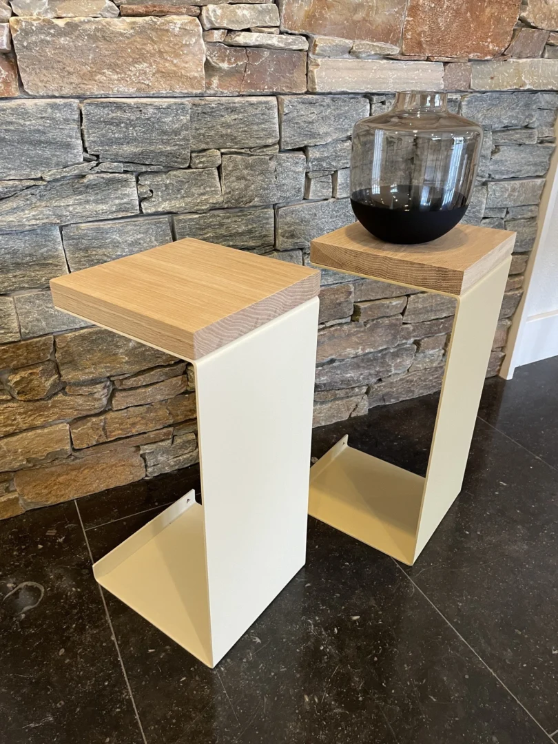 Twee Meni20 - Standaard - Zand bijzettafels met houten bladen staan op een donkere stenen vloer voor een stenen muur. Op één tafel staat een donkere glazen vaas.