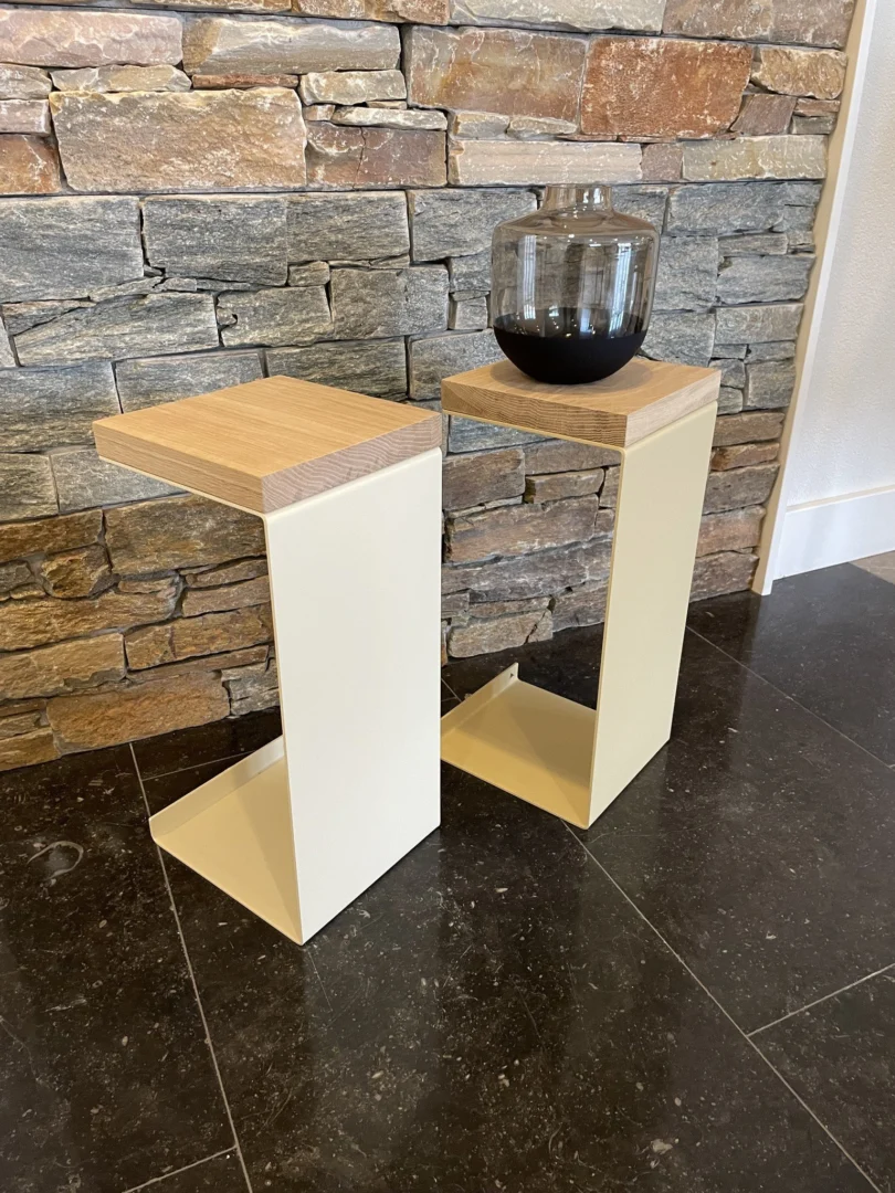 Twee Meni20 - Standaard - Zand bijzettafels met houten bladen staan op een donkere stenen vloer bij een bakstenen muur. Op één tafel staat een ronde, transparante vaas gevuld met donkere vloeistof.