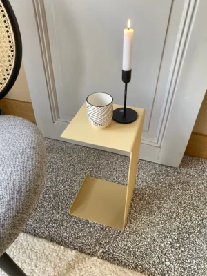 De Meni20 - Standaard - Zand, een compacte moderne bijzettafel, toont een gestreept kopje en een brandende witte kaars in een zwarte houder naast een grijze stoel met structuur en een wit gelambriseerde muur op gespikkeld tapijt.