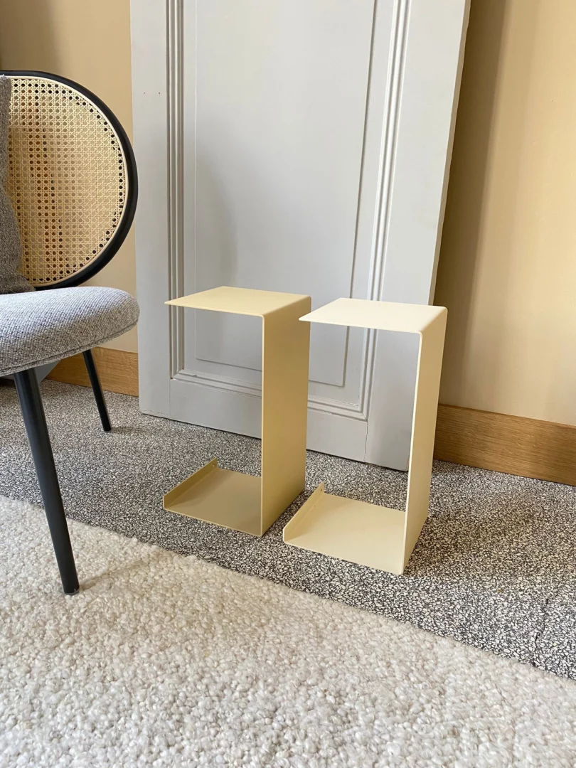 Twee Meni20 - Standaard - Zand bijzettafels met een modern, Z-vormig ontwerp staan op een vloerbedekking naast een grijs gestoffeerde stoel met een rieten rug, tegen een beige muur en witte deur.