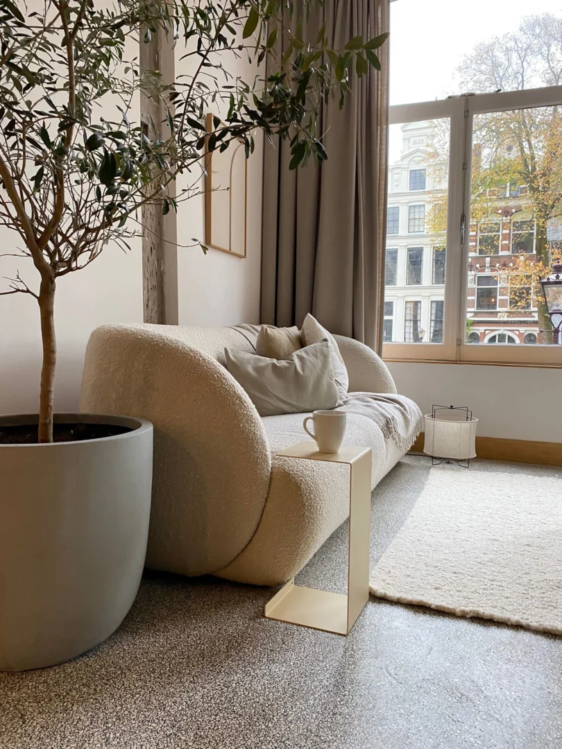 Een moderne woonkamer met de Meni20 - Standaard - Zand boucle bank, neutrale kussens, een gouden bijzettafel met een witte mok, een grote boom in pot, een licht vloerkleed, een groot raam en gebouwen in de stad die buiten te zien zijn.