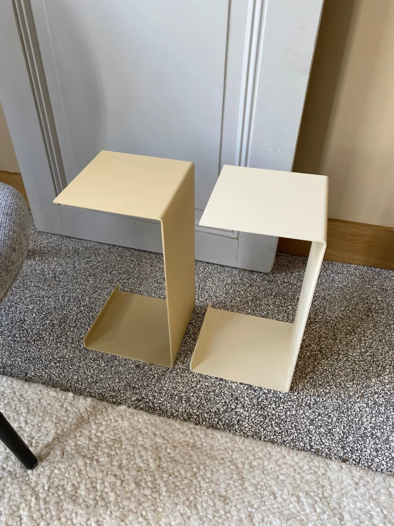 Twee Meni20 - Standaard - Zand tafels met een minimalistisch, lichtgekleurd C-vormig ontwerp staan op een tweekleurig tapijt naast een deur en een stoel.