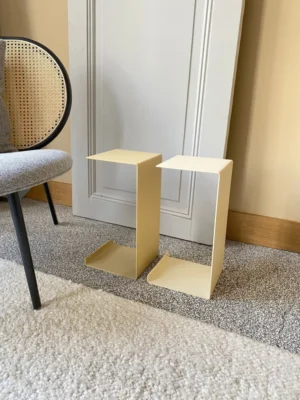 Twee Meni20 - Standaard - Crème moderne Z-vormige bijzettafels staan op een gespikkeld tapijt naast een grijs gestoffeerde stoel met een rieten rugleuning en een witte deur die tegen een beige muur leunt.