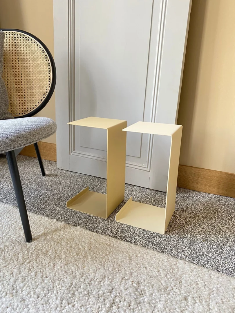 Twee Meni20 - Standaard - Crème moderne Z-vormige bijzettafels staan op een gespikkeld tapijt naast een grijs gestoffeerde stoel met een rieten rugleuning en een witte deur die tegen een beige muur leunt.