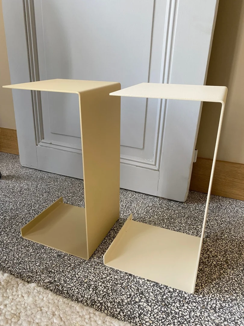 Twee Meni20 - Standaard - Crème metalen C-vormige bijzettafels met platte bladen staan op een grijs getextureerd tapijt voor een gesloten witte deur.