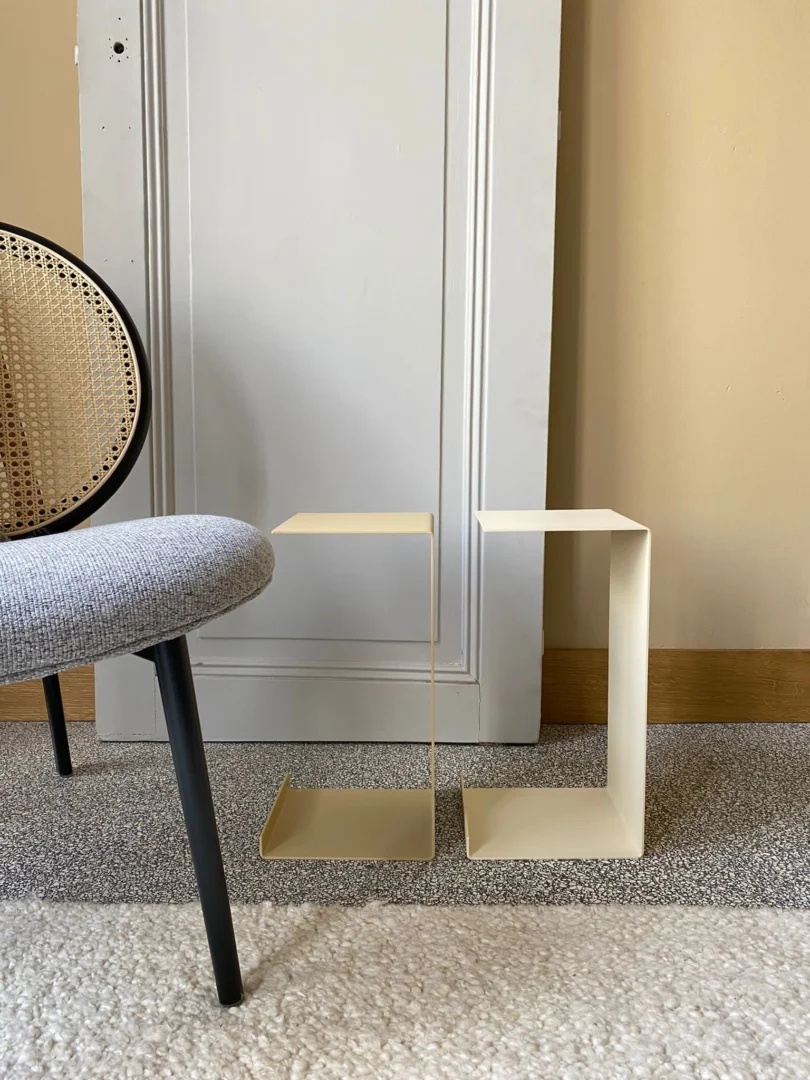 Een minimalistische kamer met een grijs gestoffeerde stoel, een gedeeltelijk zicht op een ronde rieten rugleuning en twee Meni20 - Standaard - Crème bijzettafeltjes met geometrische zigzagvormen op een tapijt met textuur bij een witte deur.