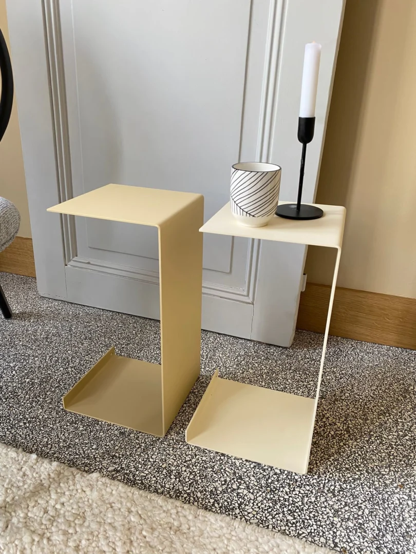 Twee crème Meni20 - Standaard metalen bijzettafeltjes met een moderne C-vorm staan op een gespikkeld tapijt. Op het ene staat een kaars in een zwarte houder en een wit gestreept kopje; het andere is leeg.