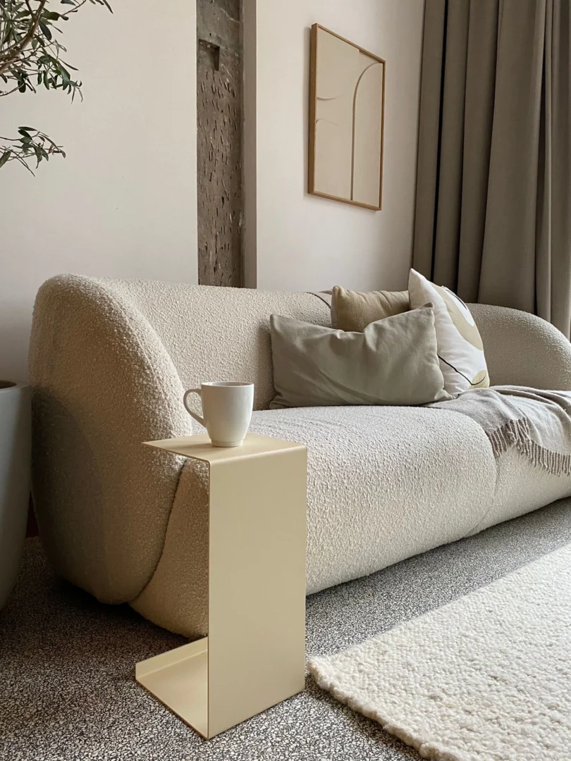 In een moderne, gezellige woonkamer staat de bank Meni20 - Standaard - Crème met neutrale kussens, een beige deken, een minimalistische crèmekleurige bijzettafel met een witte mok, abstracte kunst en zachte neutrale gordijnen voor een serene sfeer.