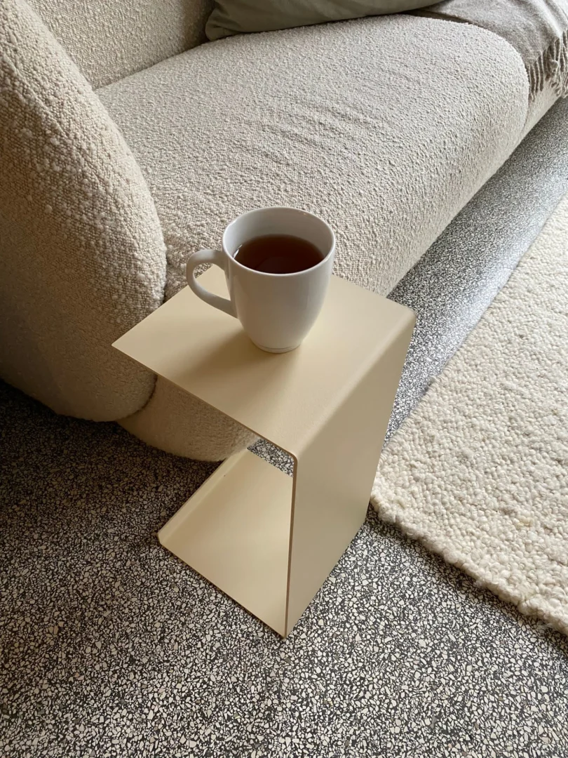 Een witte mok thee rust op de Meni20 - Standaard - Crème Z-vormige tafel naast een beige bank met textuur en een crèmekleurig tapijt op een gespikkeld grijs tapijt.