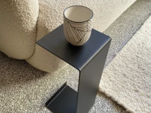 Een keramische beker met patroon rust op de Meni15 - Stalen Stand - Zwart, geplaatst naast een crèmekleurige bank met textuur en een gespikkeld tapijt op een terrazzovloer.