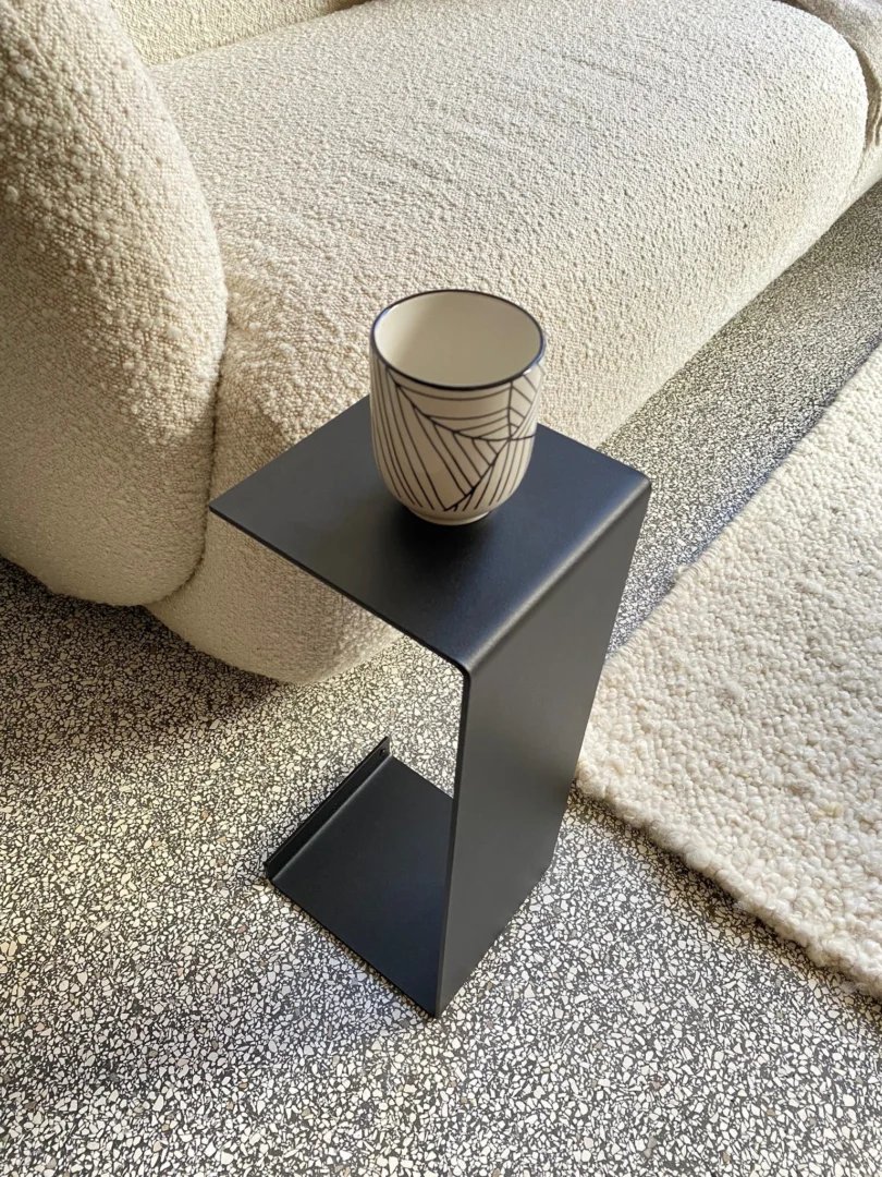 Een keramische beker met patroon rust op de Meni15 - Stalen Stand - Zwart, geplaatst naast een crèmekleurige bank met textuur en een gespikkeld tapijt op een terrazzovloer.