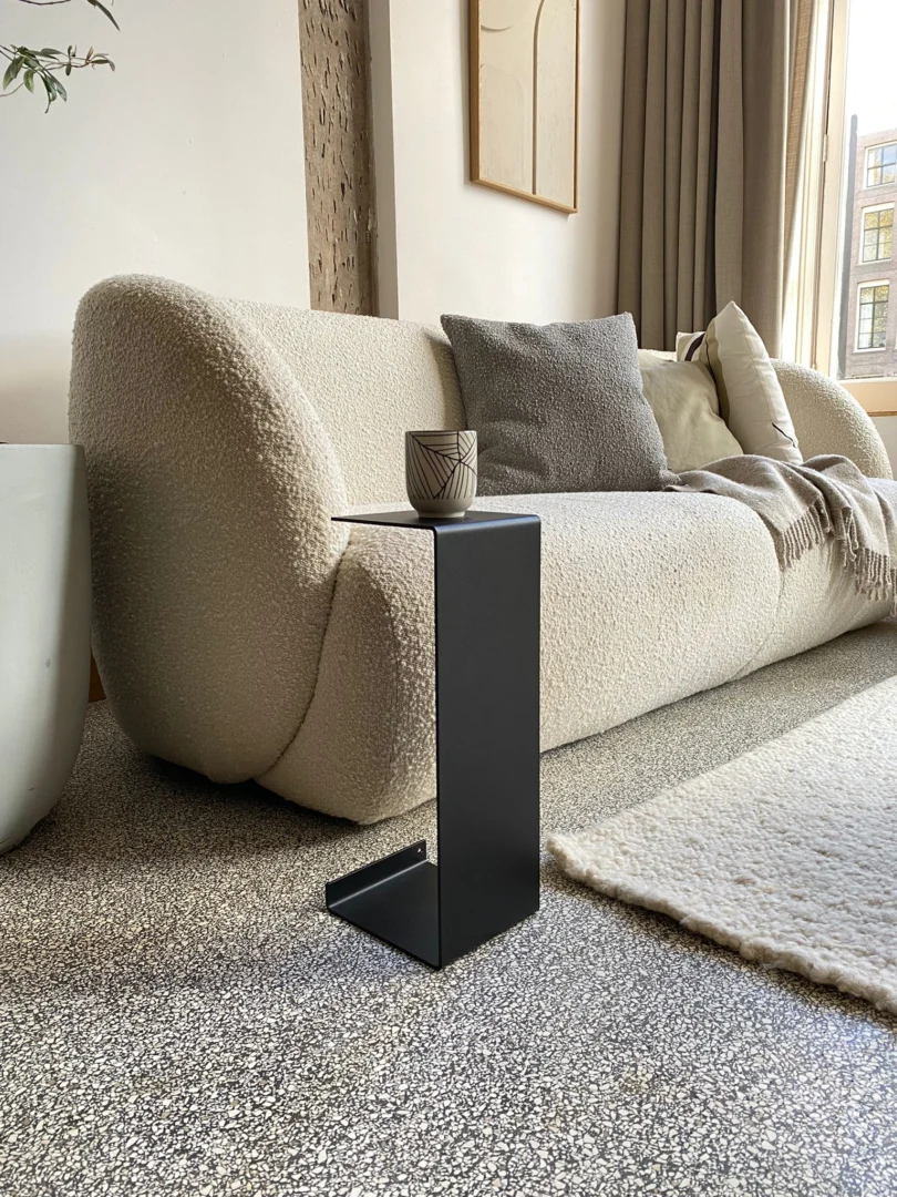 Een moderne woonkamer met een gebogen crèmekleurige bank, neutrale kussens, een vloerkleed met structuur en een Meni15 - Stalen Stand - Zwart bijzettafel met een keramische beker, omlijst door grote ramen die natuurlijk licht binnenlaten.