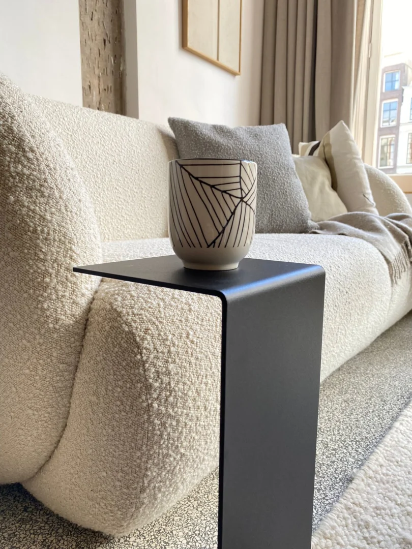 Een moderne, gezellige woonkamer met een gestructureerde witte bank, kussens en een neutrale deken, met de Meni15 - Stalen Stand - Zwart als een strak zwart metalen bijzettafeltje, plus een keramische mok met patroon bij een groot raam met beige gordijnen.
