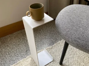 Een kleine bruine mok rust op de Meni15 - Stalen standaard - Wit, een minimalistisch wit metalen bijzettafeltje in de vorm van een omgekeerde L, naast een grijs gestoffeerde stoel met een raam en neutrale gordijnen op de achtergrond.