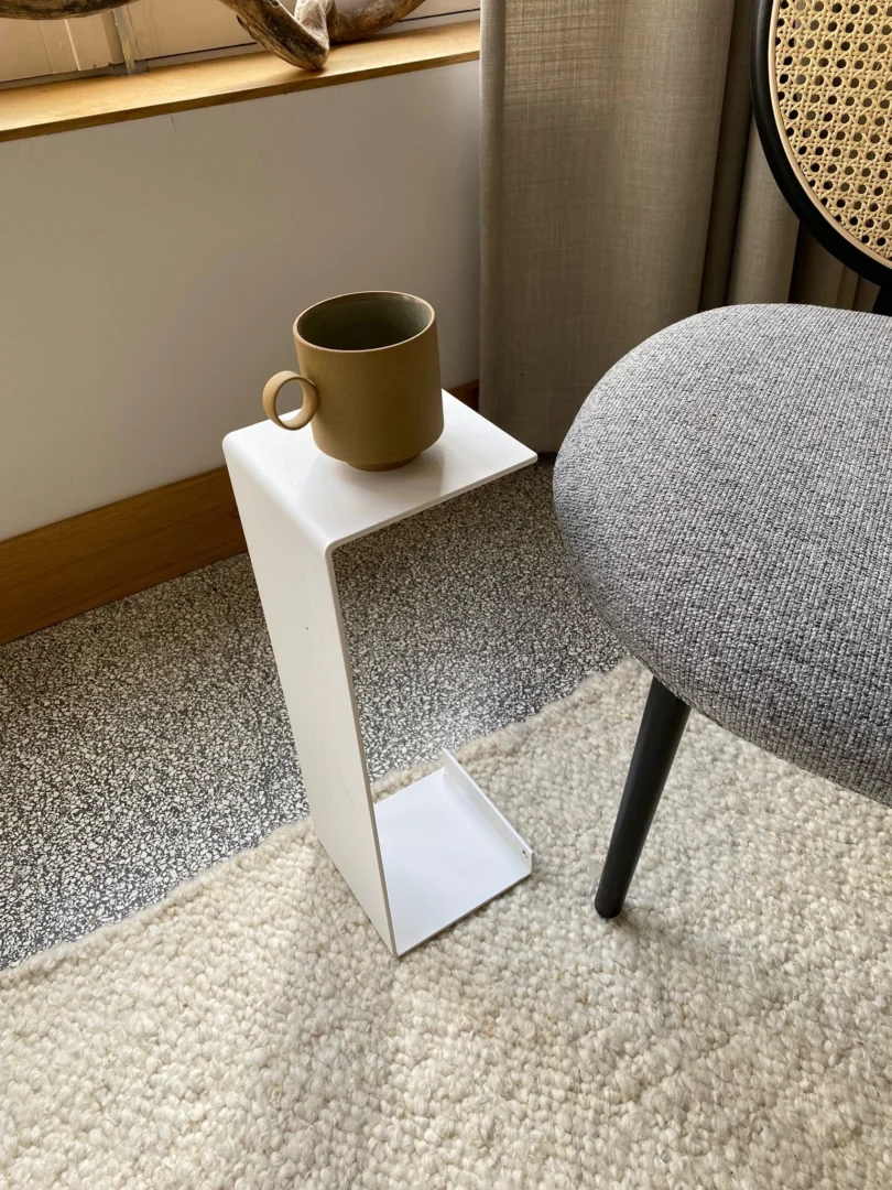 Een kleine bruine mok rust op de Meni15 - Stalen standaard - Wit, een minimalistisch wit metalen bijzettafeltje in de vorm van een omgekeerde L, naast een grijs gestoffeerde stoel met een raam en neutrale gordijnen op de achtergrond.