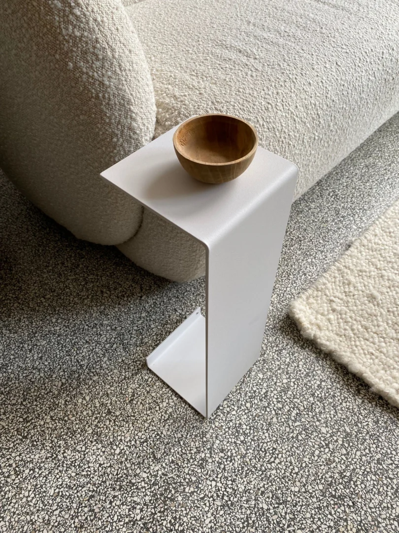 De Meni15 - Stalen standaard - Wit, een kleine, moderne witte bijzettafel met een zigzagontwerp, houdt een ronde houten schaal vast naast een lichte bank met textuur op een grijs gespikkeld tapijt, met een crèmekleurig vloerkleed ernaast.
