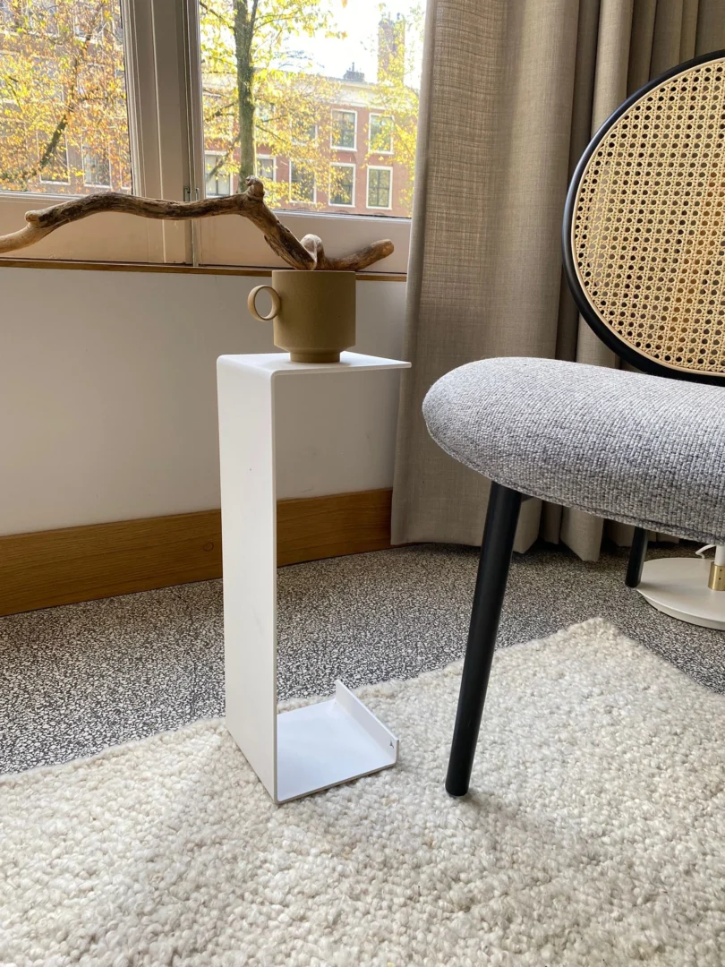 De Meni15 Stalen standaard in wit heeft een bruine mok en een decoratieve tak bij het raam met herfstbomen. Vlakbij staat een moderne stoel met een geweven rugleuning en grijs kussen op een licht getextureerd tapijt.