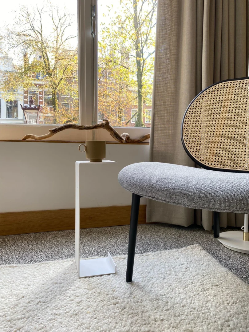 Een gezellig hoekje met een moderne grijze stoel naast de Meni15 - Stalen standaard - Wit die een beige mok vasthoudt, met beige gordijnen gedeeltelijk open en herfstbomen zichtbaar door het raam die natuurlijk licht binnenlaten.