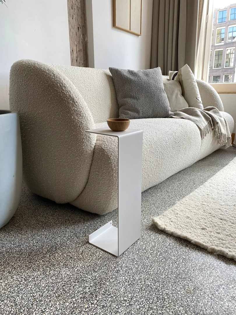 Een moderne woonkamer met een crèmekleurige boucle bank, grijze en beige kussens, een neutraal kleed, een licht vloerkleed en een kleine witte Meni15 - Stalen standaard - Wit die een houten schaal vasthoudt. Grote ramen laten natuurlijk licht binnen.