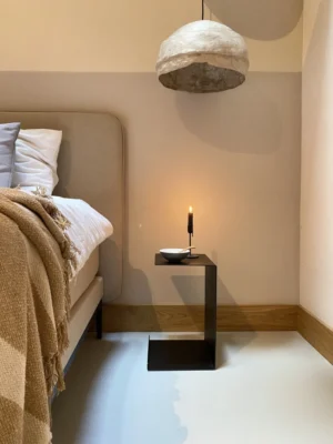Een moderne slaapkamerhoek met een beige gestoffeerd bed, een geblokte bruine deken, een minimalistisch zwart bijzettafeltje met een brandende kaars en schaal en de koepelvormige hanglamp Meni30/20 - Standaard - Zwart die erboven hangt.
